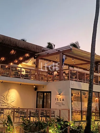 The exterior of ISSA Goa at dusk, showcasing its "Goa-terranean Luxury Shack" design. The architecture is simple yet elegant, with a focus on the river views and creating a warm, inviting atmosphere for diners.