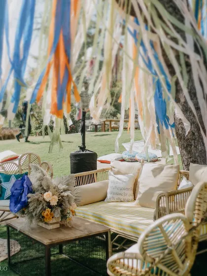 A bohemian lounge area under a tree decorated with colorful ribbons. The cane sofa and chairs, paired with a rustic coffee table, create a perfect spot to relax and soak in the festival vibes.
