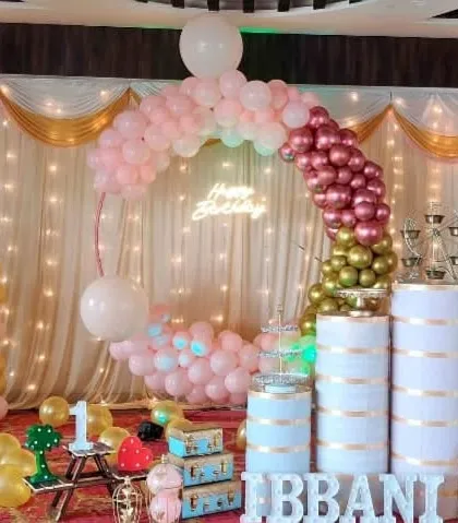 An elegant party setup with a pink and gold balloon arch, draped curtains, and stylish pedestals.