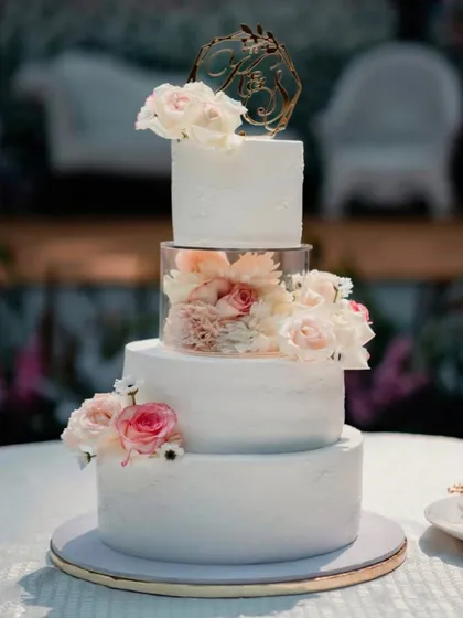 A stunning four-tier wedding cake featuring a clear spacer tier filled with fresh flowers. This design creates a beautiful floating effect, adding a modern and ethereal touch to a classic white wedding cake.