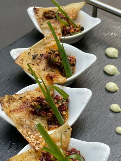 A beautifully plated appetizer featuring a crispy base topped with a savory mixture and fresh herbs. The dots of sauce on the slate platter show our attention to artistic detail.