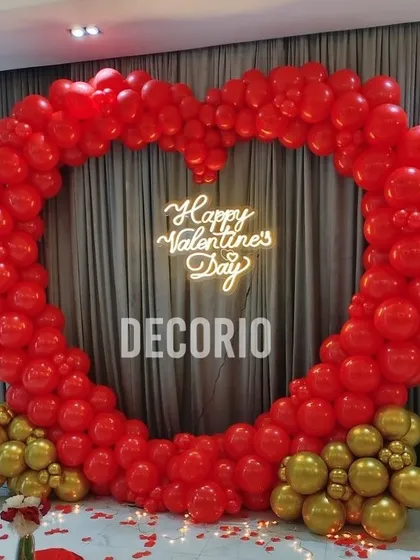A romantic Valentine's Day photo booth designed with a large heart-shaped balloon arch in red and gold. The "Happy Valentine's Day" neon sign adds a warm glow, making it an ideal backdrop for couples.