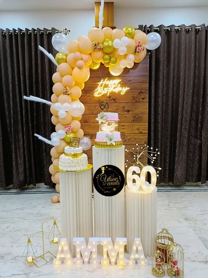 An elegant 60th birthday celebration for 'Amma'. The decor features a pastel balloon arch against a wooden backdrop, with two cakes and marquee numbers.