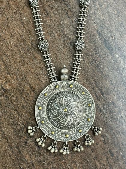 A large, circular oxidised silver pendant with a shield-like design. The chain is also detailed with small floral links and the pendant has ghungroo drops.