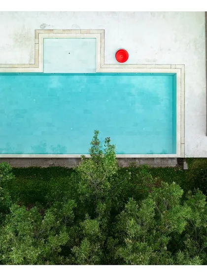 An aerial view of a minimalist swimming pool, with a single red accent chair providing a pop of color. The clean lines of the pool and surrounding white paving create a serene and modern aesthetic.