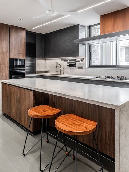 The island in this Ahmedabad kitchen provides a casual seating area, with warm wooden stools complementing the walnut cabinetry and contrasting with the dark island base.