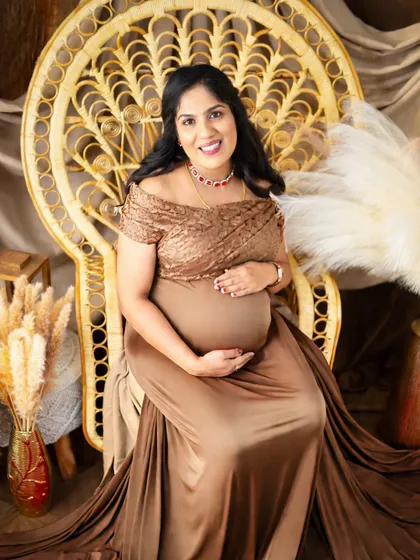 A portrait of pure elegance. The mom-to-be is seated on a peacock wicker chair, wearing a beautiful brown satin gown that drapes gracefully around her.
