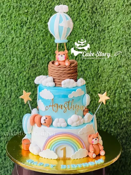 A dreamy three-tier cake for a first birthday. The theme features a teddy bear in a hot air balloon, floating among the clouds and a rainbow, creating a beautiful story.
