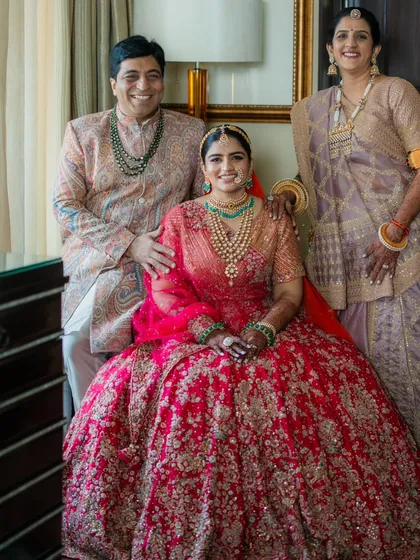 A cherished portrait of the bride with her parents. The father's paisley sherwani and the mother's muted gold saree perfectly complement each other and the bride's vibrant red, creating a picture of coordinated elegance.