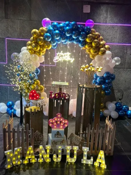 A grand birthday setup with a rustic touch. This decoration includes a blue and gold balloon arch, a picket fence, light-up letters, and various props to create a unique party corner.