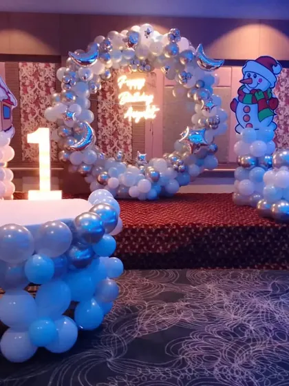 A winter wonderland theme on a stage in a banquet hall. The setup includes a silver and white balloon ring, snowman and house cutouts, and a light-up number '1'.