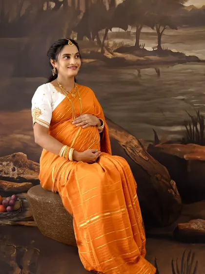 A solo portrait of a mom-to-be in an orange saree, seated in a painted natural landscape, looking peaceful and radiant.