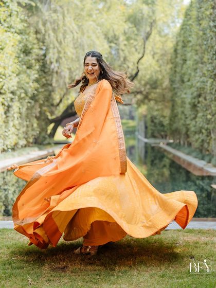 A bride twirls in her vibrant orange lehenga, her happiness radiating in the beautiful garden setting of her Haldi function.