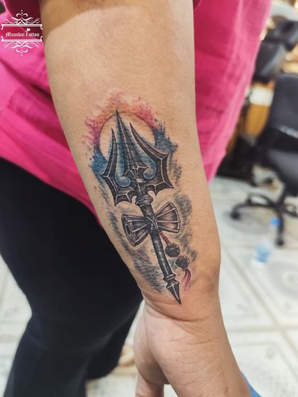 A vibrant watercolor-style Trishul tattoo. The blend of red and blue behind the black and grey trident gives the design a dynamic and energetic feel.