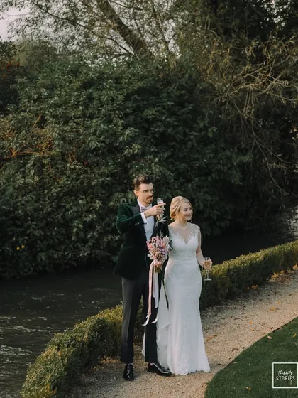 The couple sharing a toast by the river at their Bibury wedding. This candid moment captures the celebratory and serene atmosphere of their special day.