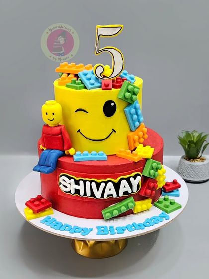 A fun and colorful two-tier Lego cake, with a winking emoji face and edible Lego blocks tumbling down the sides.