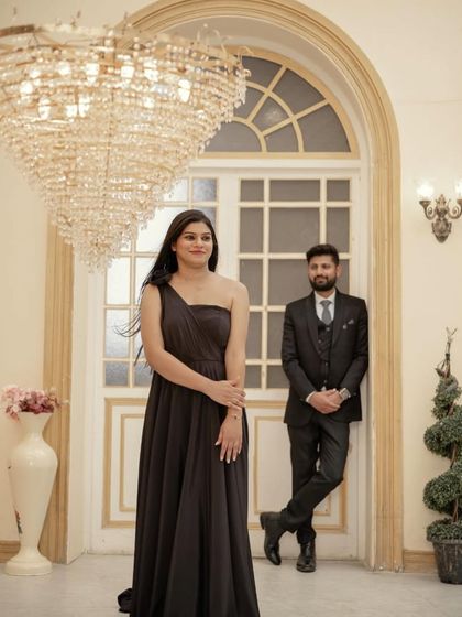 A sophisticated pre-wedding shot with the couple in a grand room. The woman wears a simple yet elegant black one-shoulder gown.