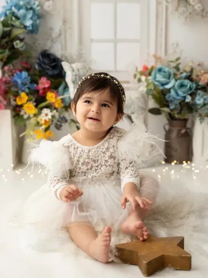 A smiling baby girl in an elegant white lace dress, surrounded by a colorful floral setup. This is a beautiful option for a first birthday photoshoot.