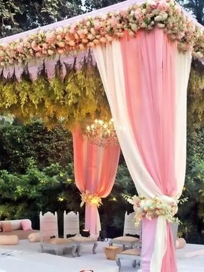 A beautiful pastel-themed outdoor mandap. The soft pink and white drapes, combined with a roof of delicate flowers and greenery, create a light, airy, and romantic setting for a daytime wedding.