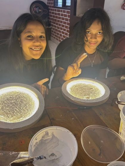 Two students smiling as they work on their moon lamps, with the lights on to help them see the texture they are creating.