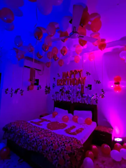 A romantic birthday room decoration designed for a special surprise. The room is filled with red and white balloons on the ceiling and floor, with 'Happy Birthday' foil balloons over the bed, which is decorated with flower petals. Moody blue and pink lighting sets a perfect ambiance.