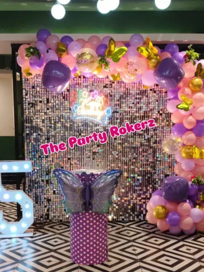 A vibrant butterfly theme using a silver sequin wall and a balloon arch in shades of purple and pink. The decor is completed with a butterfly-themed cake stand and a large marquee number '5'.