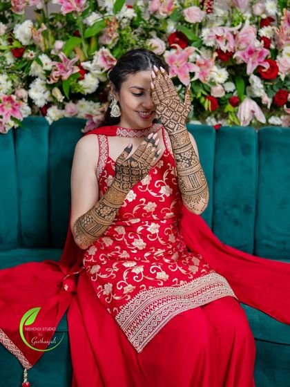 A happy bride posing against a beautiful floral backdrop, her intricate mehendi design adding to her overall look.