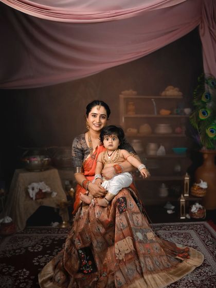 A beautiful, painterly portrait of a mother and her baby in a traditional Krishna Janmashtami setup. The rich colors and fabrics create a stunning visual.
