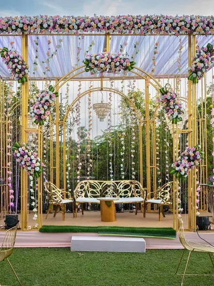 A chic and modern stage setup for a Mehendi or Sangeet. The gold frame structure is decorated with pastel florals and hanging crystals, with a memory wall of photos on the side.