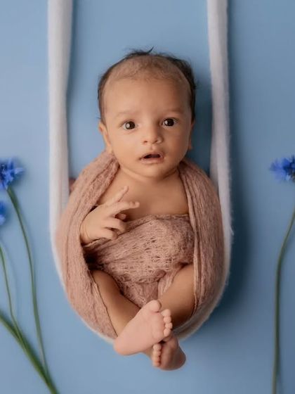 An alert newborn with wide eyes is wrapped in a brown swaddle, sitting in a soft white swing against a blue background with flowers.