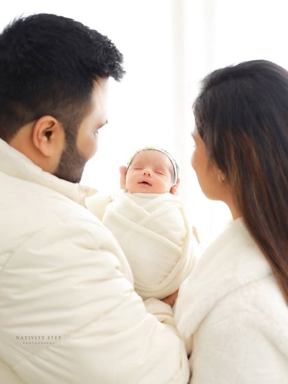 A family portrait with a clean, high-key look. The parents are dressed in white, matching the baby's swaddle, creating a cohesive and angelic family photo.