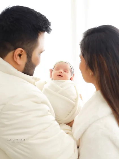 A family portrait with a clean, high-key look. The parents are dressed in white, matching the baby's swaddle, creating a cohesive and angelic family photo.