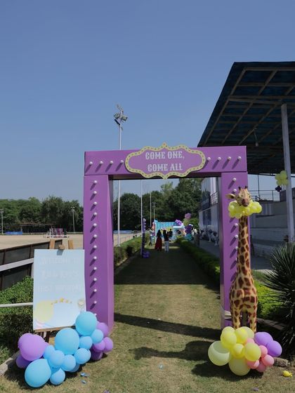A grand entrance archway for an outdoor carnival, with a "Come One, Come All" sign. A balloon-adorned giraffe prop adds a playful touch to the welcome area.