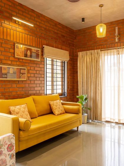 The living room of the completed Akshaynagar duplex, featuring a bright yellow sofa against an exposed brick wall. The space is finished with warm tones and Kota stone flooring, creating a cheerful and earthy atmosphere.