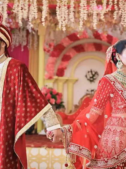 A regal portrait of the couple holding hands, showcasing their coordinated red and white wedding outfits against a decorated backdrop.