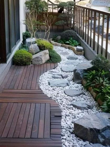 This balcony is transformed into a miniature Japanese-style garden. It features a combination of wooden decking and a dry riverbed of white pebbles, complete with rocks and bonsai-like plants.