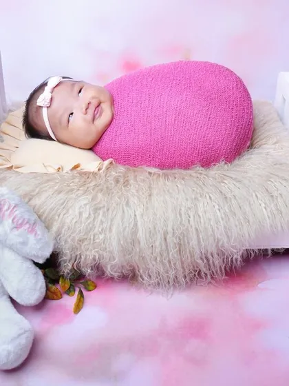 This wide-eyed baby girl is all smiles while lying in a miniature bed, accompanied by a soft toy bunny. The pink floral backdrop adds to the cheerful atmosphere.