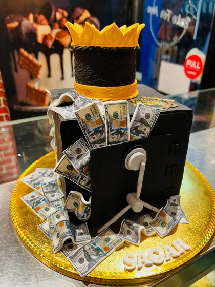 A creative money-themed cake designed as a safe overflowing with edible dollar bills, topped with a golden crown.