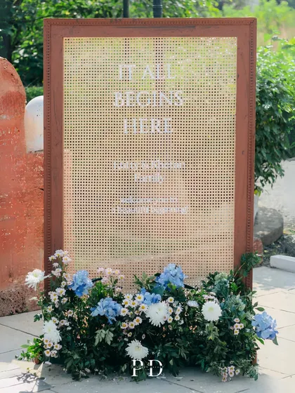 A unique welcome sign for a boho-chic Mehendi, using a rattan screen and a bed of colorful flowers. It sets a warm, natural, and stylish tone.