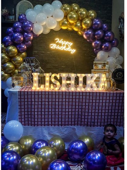 A sweet first birthday setup for Lishika right at home. A purple and gold balloon arch frames a table with her name in light-up letters, creating a perfect little photo spot for her and the family.