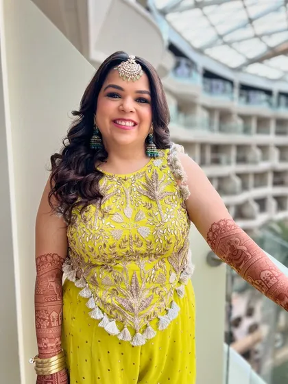 A happy client showing off her Mehendi look. The soft, full curls frame her face perfectly, paired with fresh makeup and a statement maang tikka.