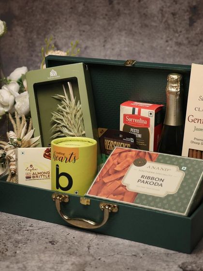 A sophisticated green trunk hamper for Diwali. It's packed with premium items like non-alcoholic champagne, artisanal pakodas, and jasmine incense, making it a truly luxurious gift.