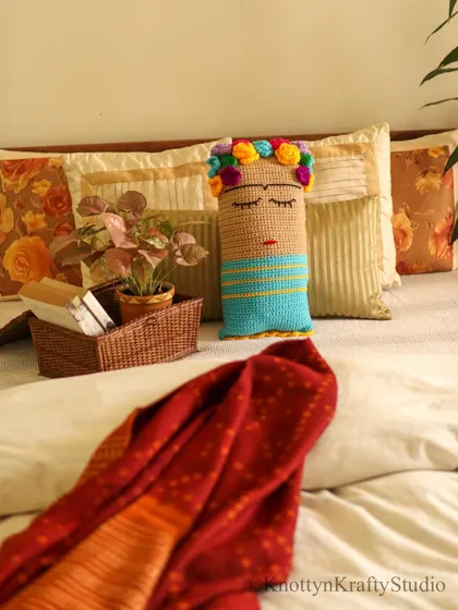 The Frida Kahlo cushion styled on a bed with a warm red throw. This view shows how it can be part of a larger, layered look to create a comfortable and inviting bedroom.