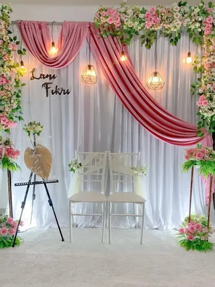 An elegant and romantic engagement stage decoration for an indoor ceremony. This setup includes a white fabric backdrop with gracefully draped pink velvet, framed by lush pink and white floral arrangements and illuminated by warm hanging pendant lights.