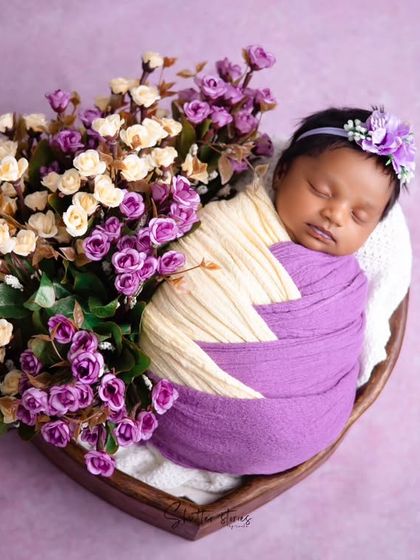 A newborn girl wrapped in purple and cream, nestled in a heart bowl with a bouquet of matching purple and white flowers.