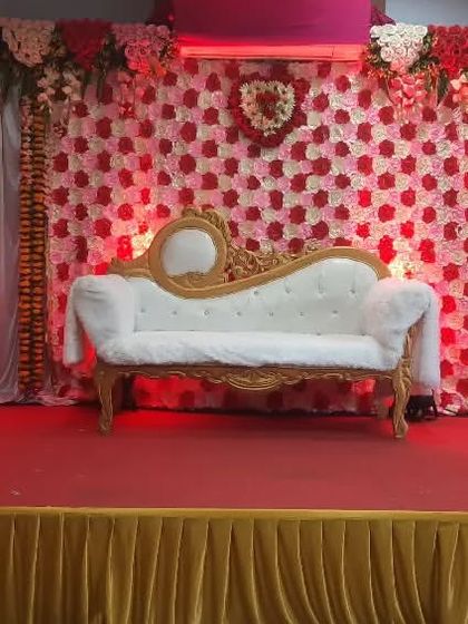 This reception stage features a stunning flower wall backdrop made of red, pink, and white roses. The design is complemented by a classic white chaise lounge and warm uplighting, creating a romantic and luxurious feel.