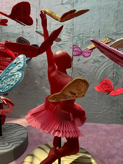A detail of a red dancer sculpture from the 'We/Us' installation. The figure's skirt is made of intricately folded paper, and it is surrounded by a swarm of colorful butterflies.