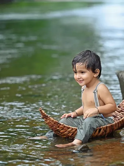 A serene portrait of the baby sitting in a wicker boat by the water's edge.
