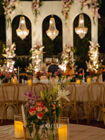 A beautifully blurred shot of the elegant outdoor dinner in Jaipur. The focus is on the candlelit floral centerpiece in the foreground, with the chandelier-adorned stage creating a magical backdrop.