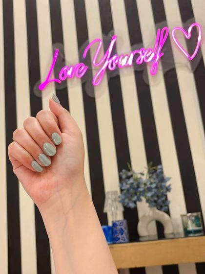 A full view of the hand with sage green holochrome nails against our salon's "Love Yourself" sign.